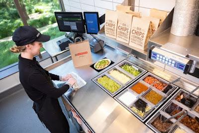 Chipotle burrito bowl being raised from automated makeline while employee assembles a burrito.