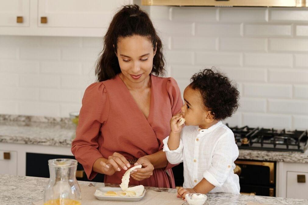 A young black mother standing next to her child in the kitchen and enjoying a healthy snack together.