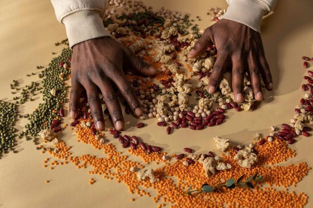An image of various grains, legumes and soy protein as a demonstration of clean vegan protein sources. The dried legumes etc are being spread around on a table by a pair of well-manicured black hands.
