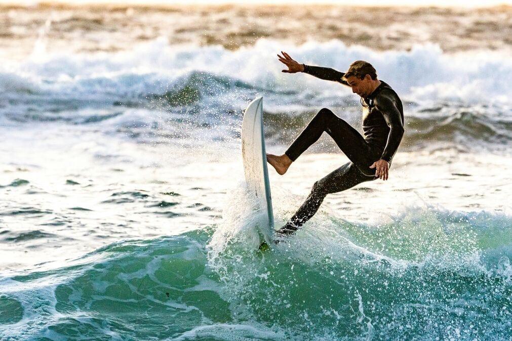An athletioc image of a male surfer battling a wave with their surfboard vertical in the water. They are wearing a wetsuit and look physically under duress but very fit.