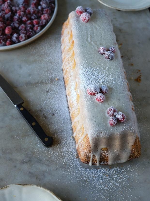 Full, unsliced cranberry cake with vanilla icing and powdered sugar decorated with sugared cranberries on marble