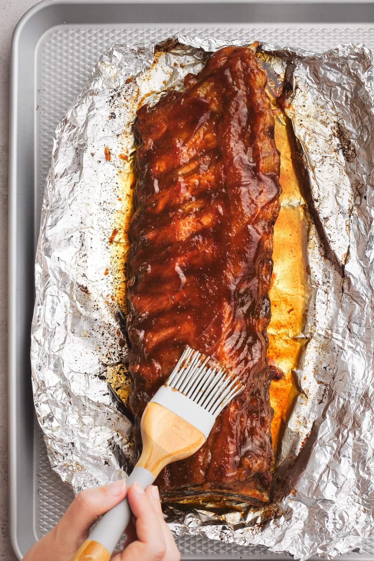 woman's hand brushing bbq sauce on ribs in foil on baking sheet
