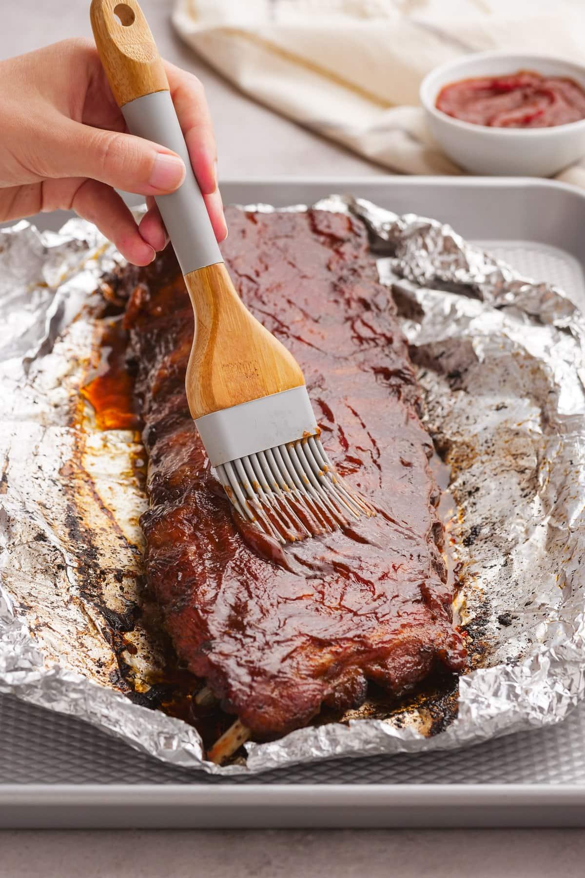 woman's hand brushing bbq sauce on ribs in foil on baking sheet