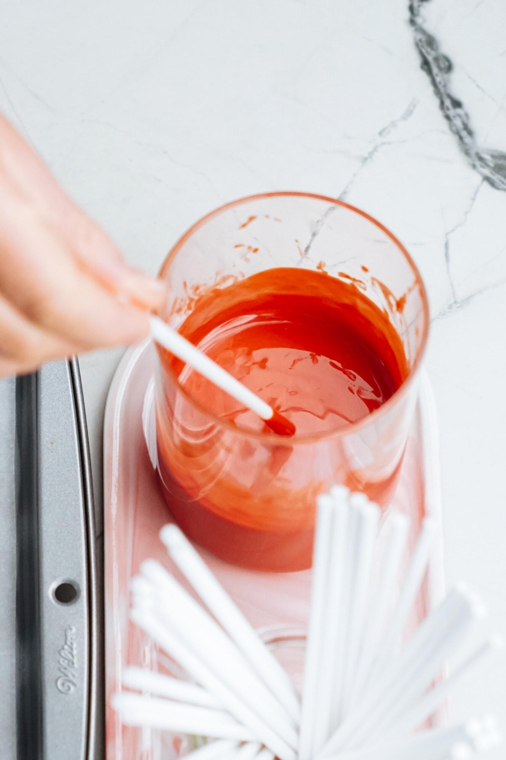 womans hand dipping the end of a lollipop stick in melted chocolate 