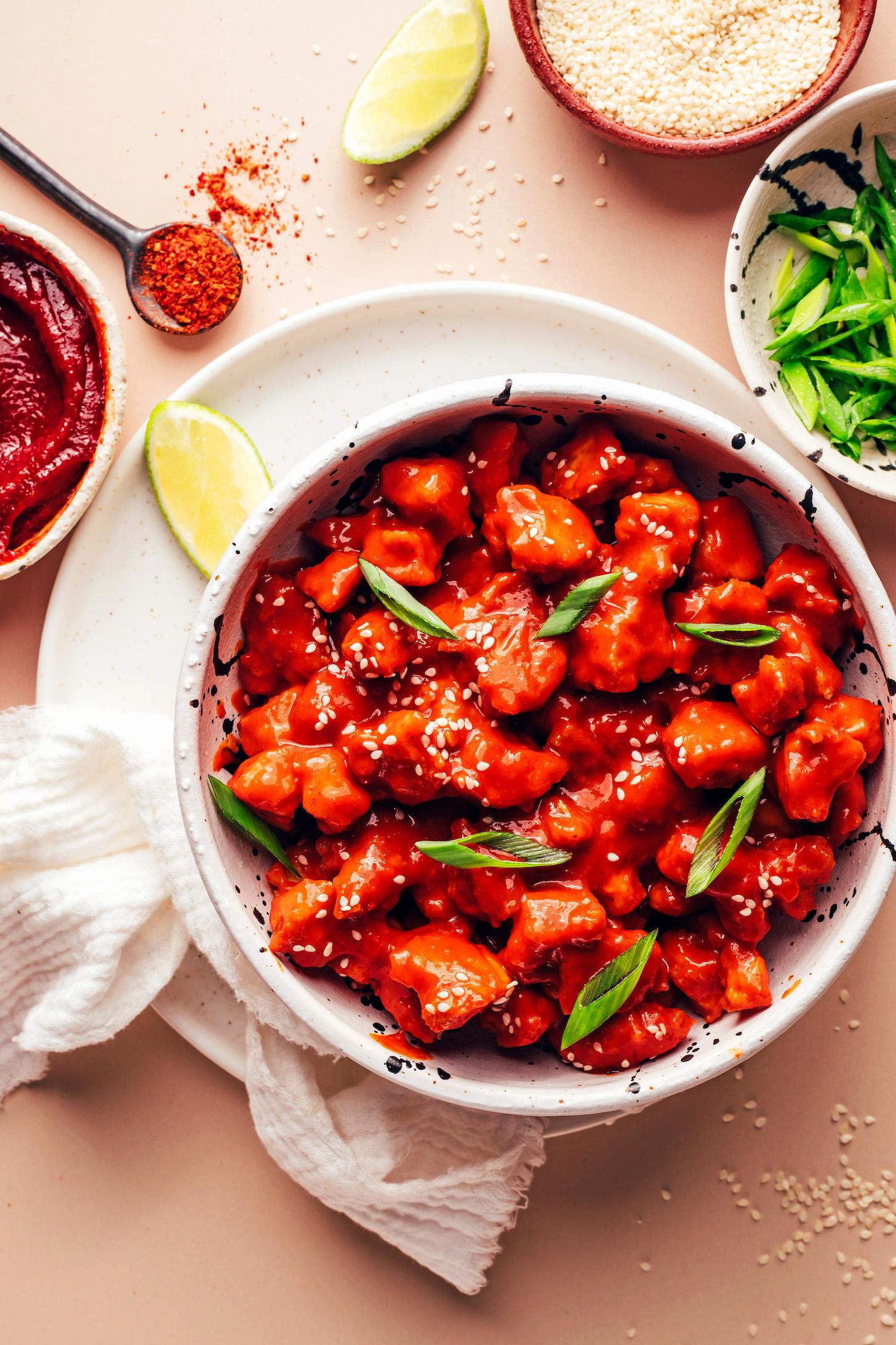 Green onions, sesame seeds, lime wedges, cayenne, and gochujang next to a bowl of crispy baked gochujang tofu