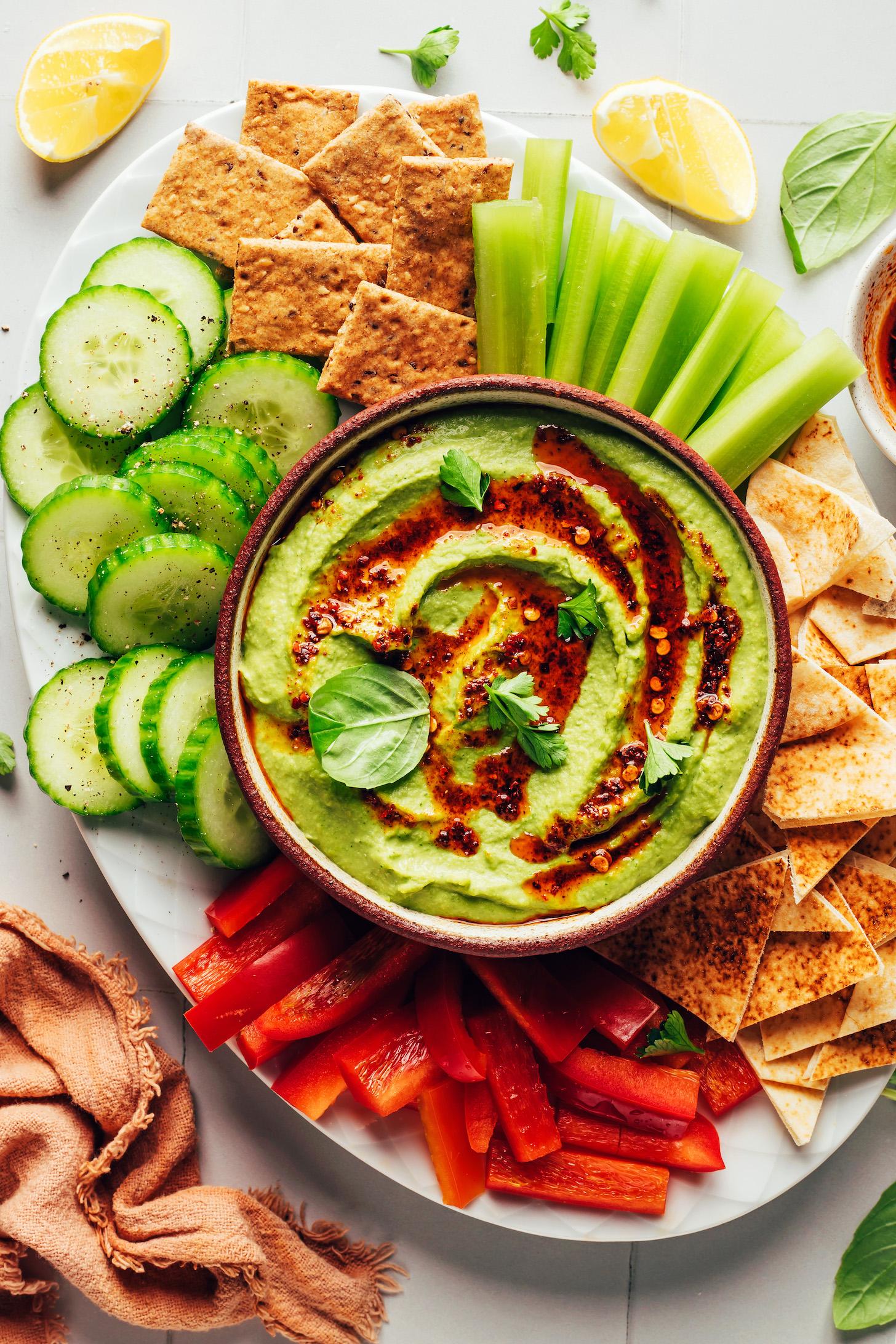 Green goddess hummus in a bowl with cucumber, crackers, celery, flatbread, and bell pepper around it