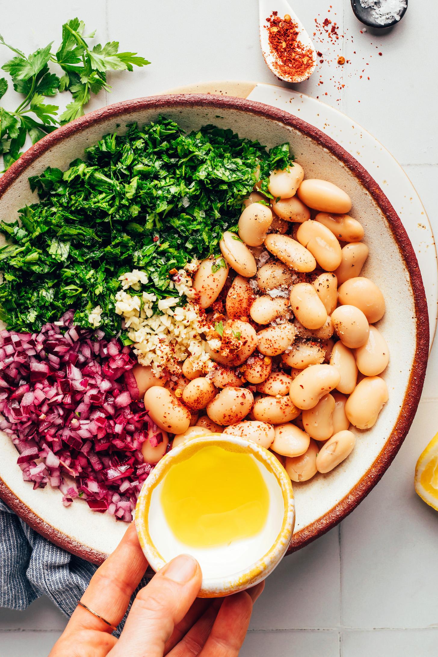 Pouring olive oil over white beans, garlic, parsley, red pepper flakes, and red onion