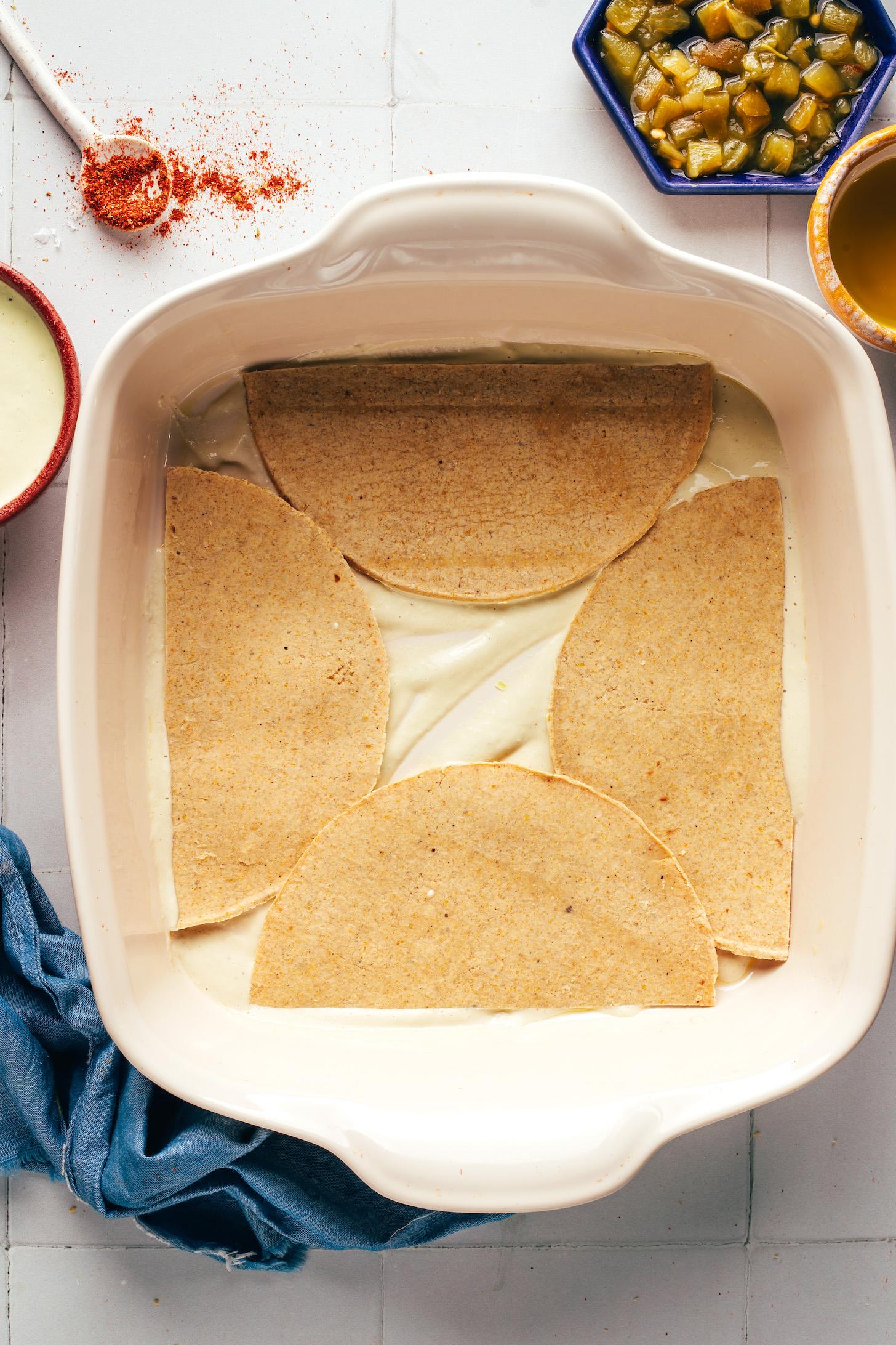 Halved corn tortillas arranged on the bottom of a casserole dish over a layer of creamy cashew sauce