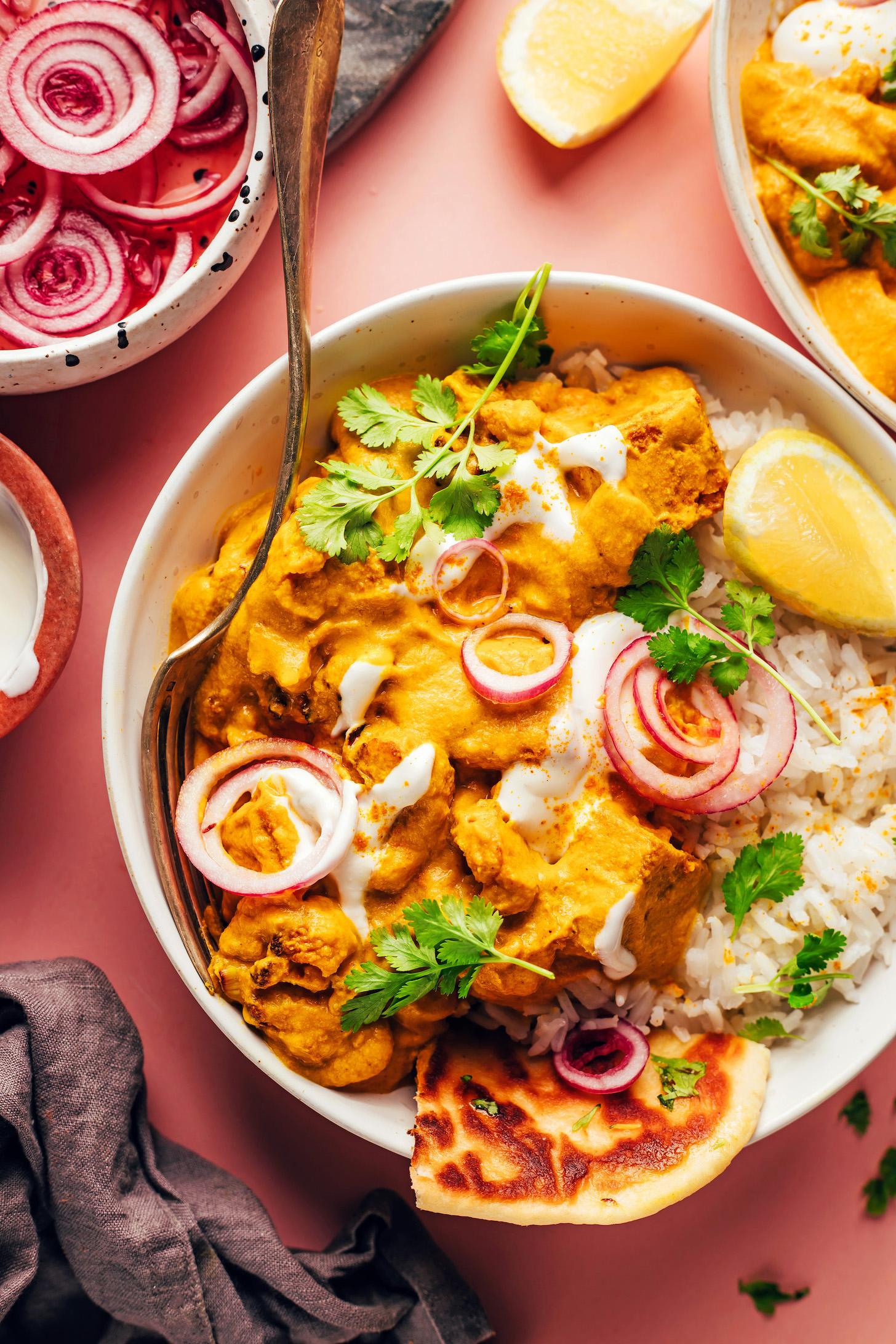 Bowl of vegan tofu cauliflower korma curry with naan, rice, lemon, cilantro, coconut yogurt, and pickled onions