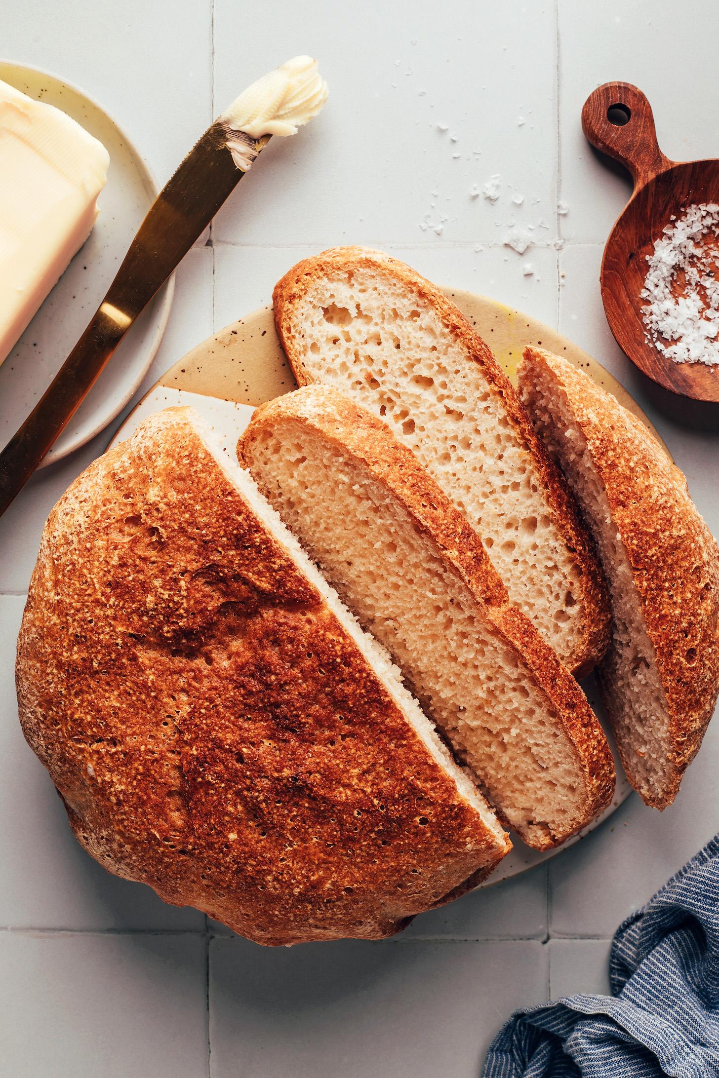 Flaky salt and butter next to a partially sliced loaf of crusty gluten-free artisan bread