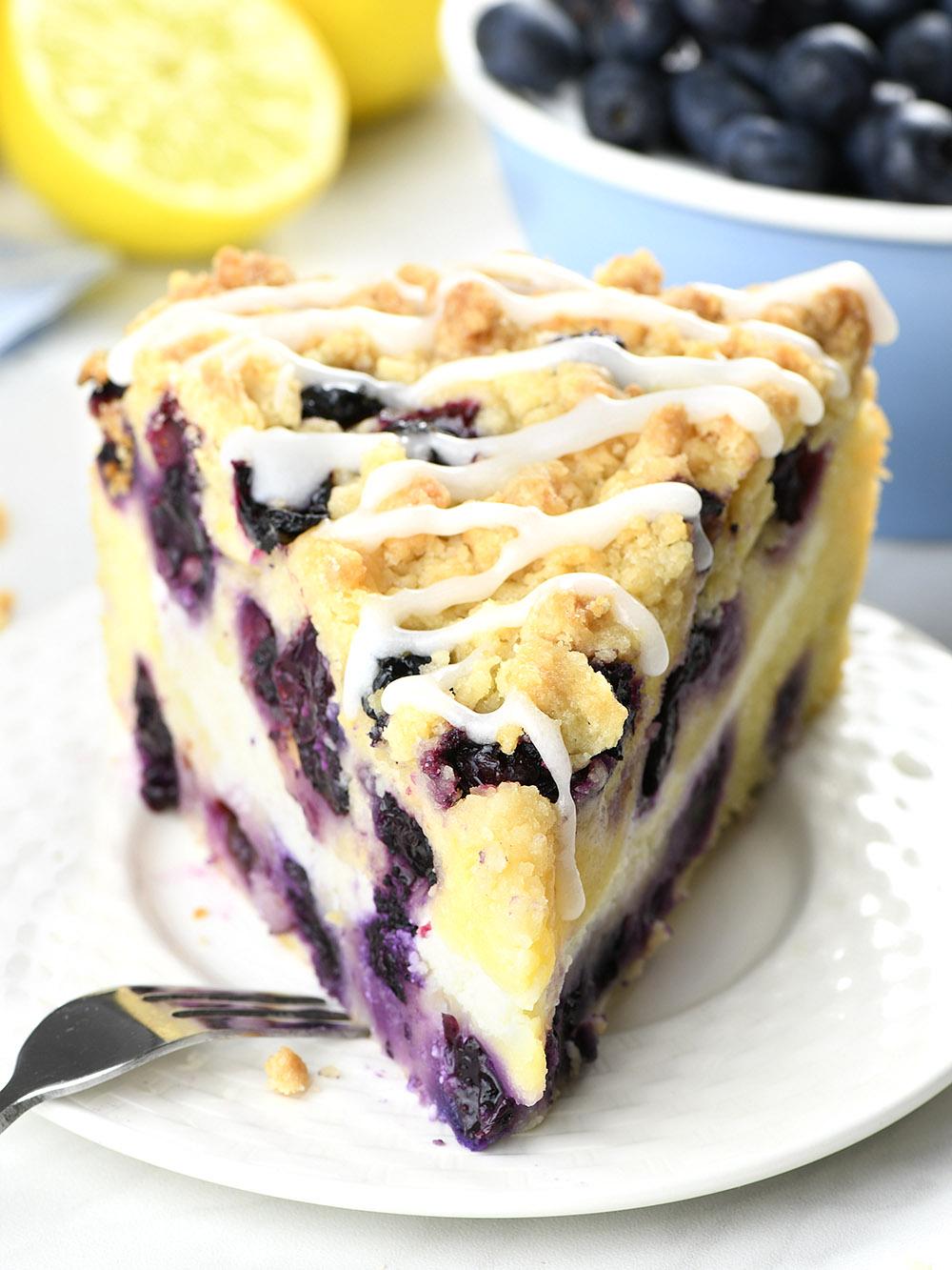 This image features a tantalizing slice of Lemon Blueberry Coffee Cake positioned front and center on a pristine white plate. The cake itself is a golden-brown hue, with a dense, moist texture visible from the cut side. Pockets of vibrant blueberries are embedded throughout, providing a lovely color contrast. The top of the cake boasts a sugary crumble topping, while a tangy lemon glaze zigzags across it, adding a glossy finish. In the soft-focus background, fresh, bright yellow lemons and plump, ripe blueberries are scattered, hinting at the natural, fruity flavors incorporated in the cake. Overall, the photo invokes a sense of freshness and indulgence.