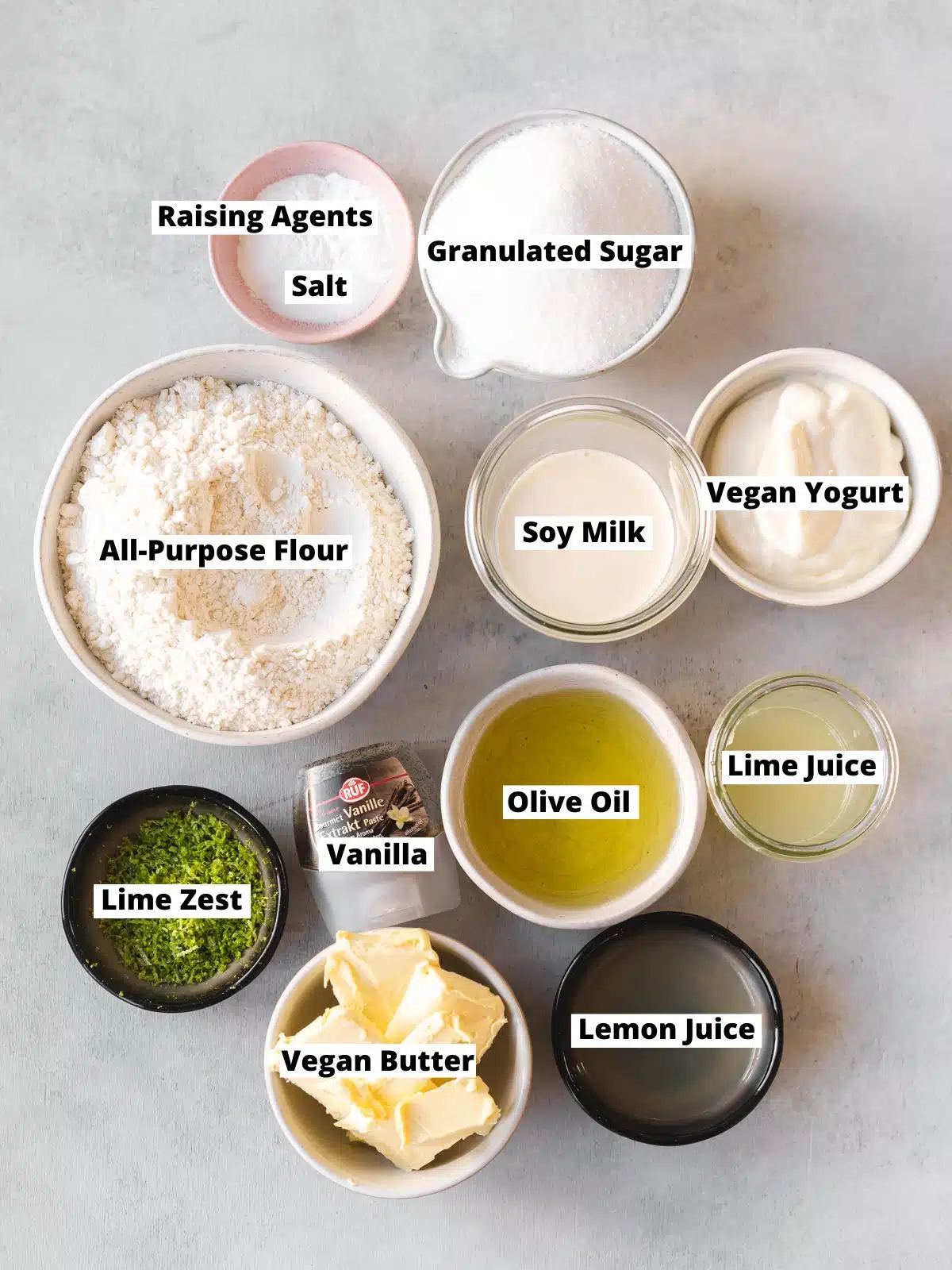 Overhead shot of ingredients needed to make easy vegan key lime cake measured out into bowls on a grey table with text overlay.