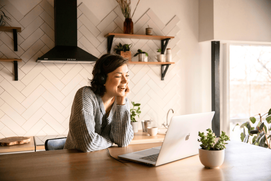 Image of a woman in a brightly lit kitchen with a laptop on an island counter, smiling at the screen.