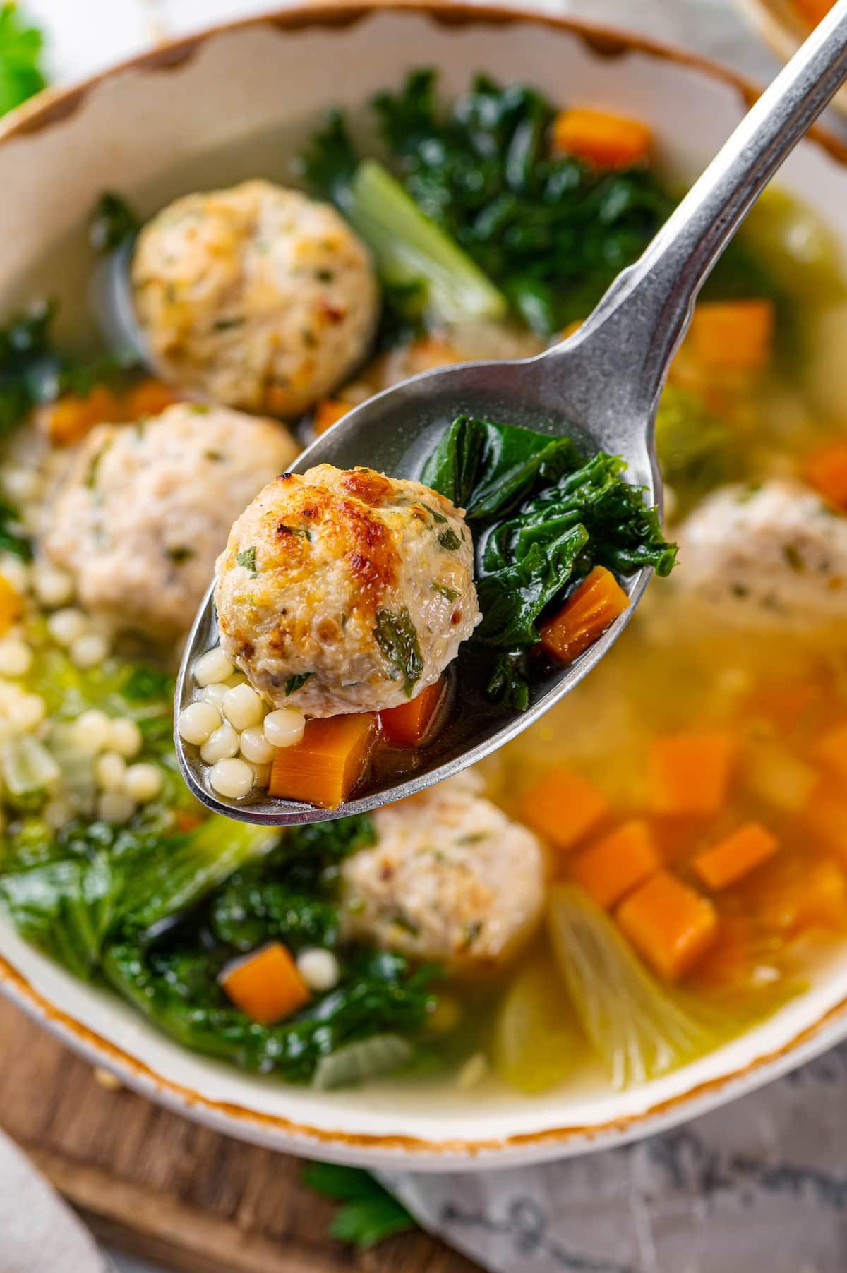 A meatball on a spoon above a bowl of Italian wedding soup.